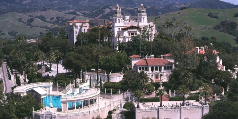 A panoramic view of Hearst Castle perched atop the San Simeon hills, framed by blue skies and lush greenery."
