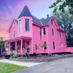 Exterior view of the Barbiecore Mansion, showcasing its vibrant pink facade and charming architecture, surrounded by lush greenery.